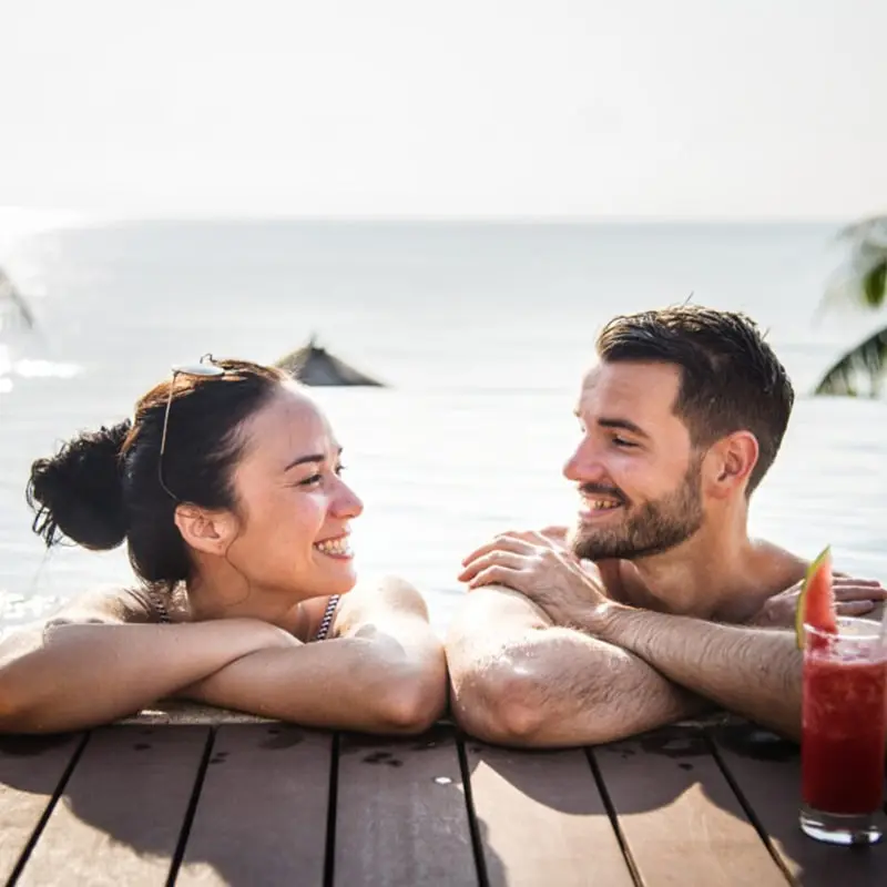 Couple smiles at each other as they rest their elbows on the dock on their honeymoon