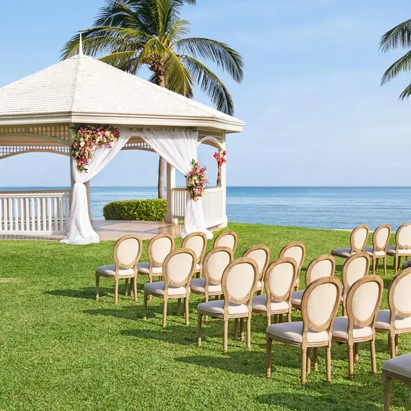 A white gazebo on grass with the ocean in the background and elegant white chairs lined up on the grass.