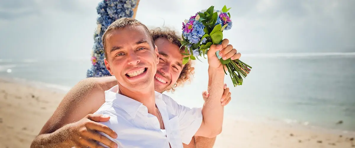 Gay couple holding a wedding bouquet and smiling on the beach