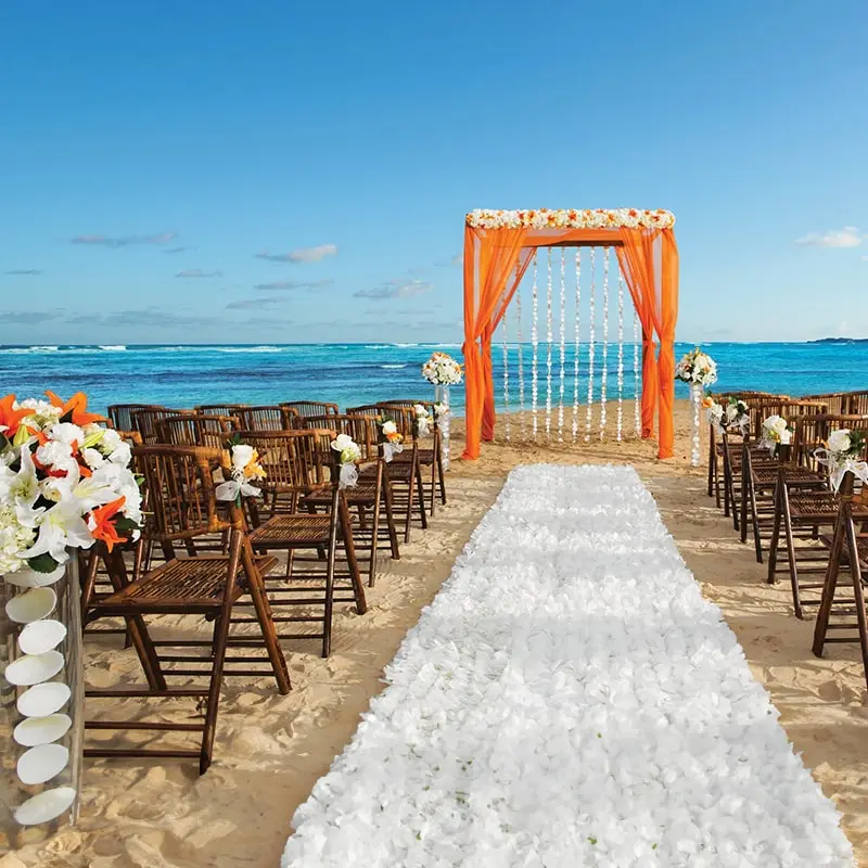 An altar on the beach is draped in orange cloth, with a white flower aisle leading towards it.