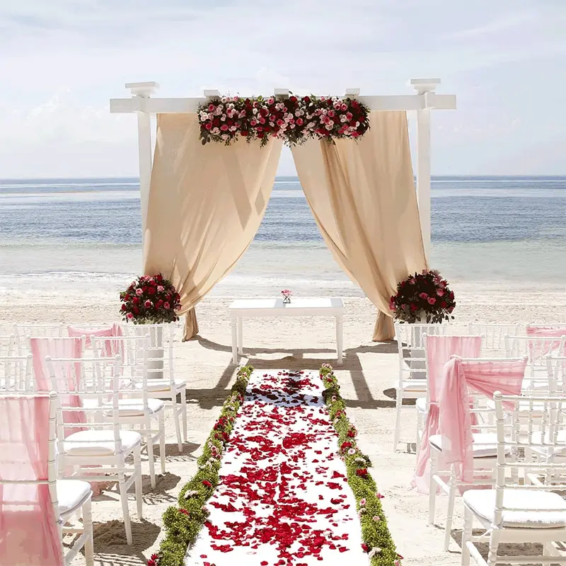 A cloth-draped altar is on a white sand beach with petals strewn across the aisle.