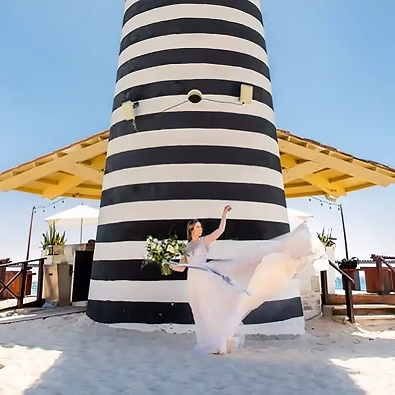 A bride's dress is blowing in the wind as she stands in front of a striped building on a white sand beach.