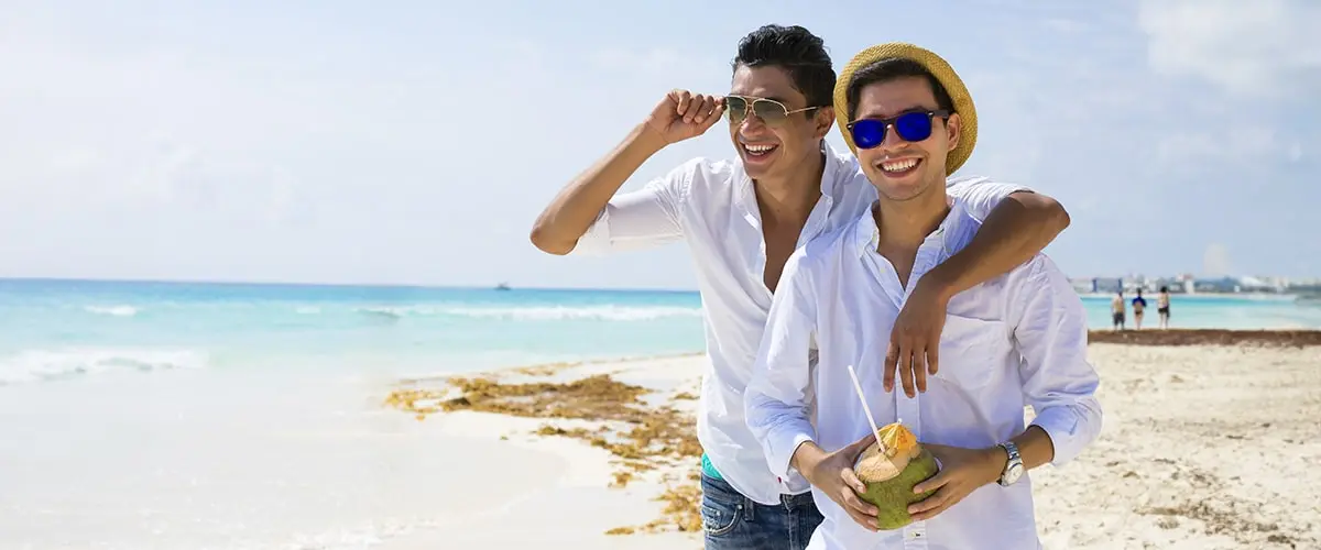 A gay couple smiling on a beach in Jamaica, one holds a coconut