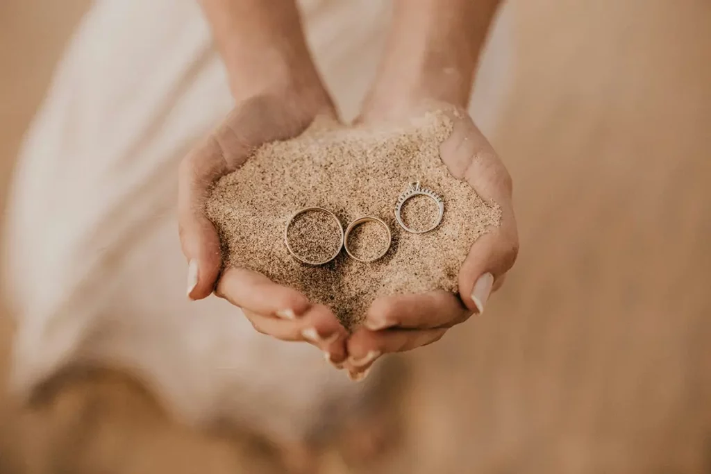 A bride holds her rings on a pile of sand in her hands on the beach