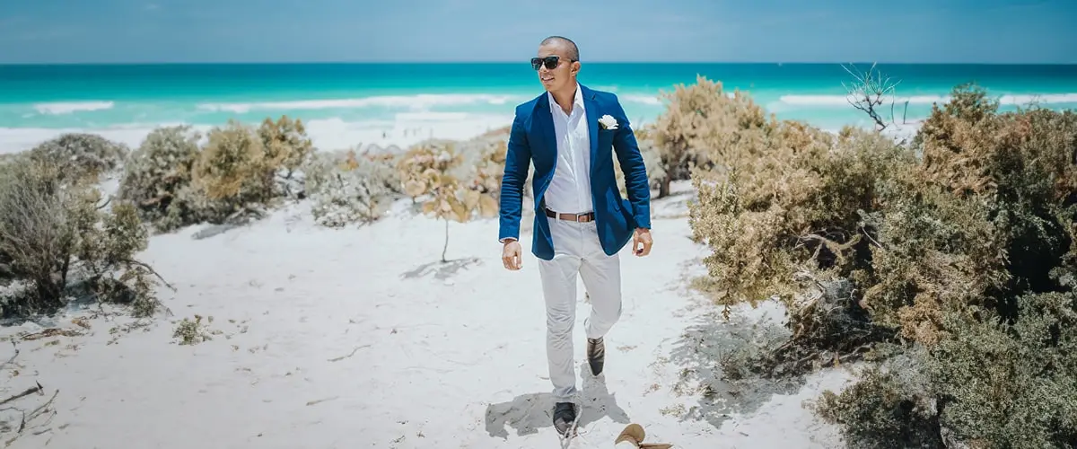 Fashion trends in destination weddings show up on the beach - a groom wears a relaxed suit with linen pants and a tropical boutonnière 